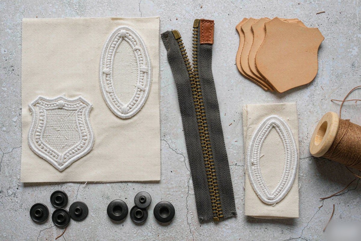 Flat lay of workwear branding components including an embroidered chest patch, woven neck label, matte black snaps, brass zipper with leather pull and leather patch blanks