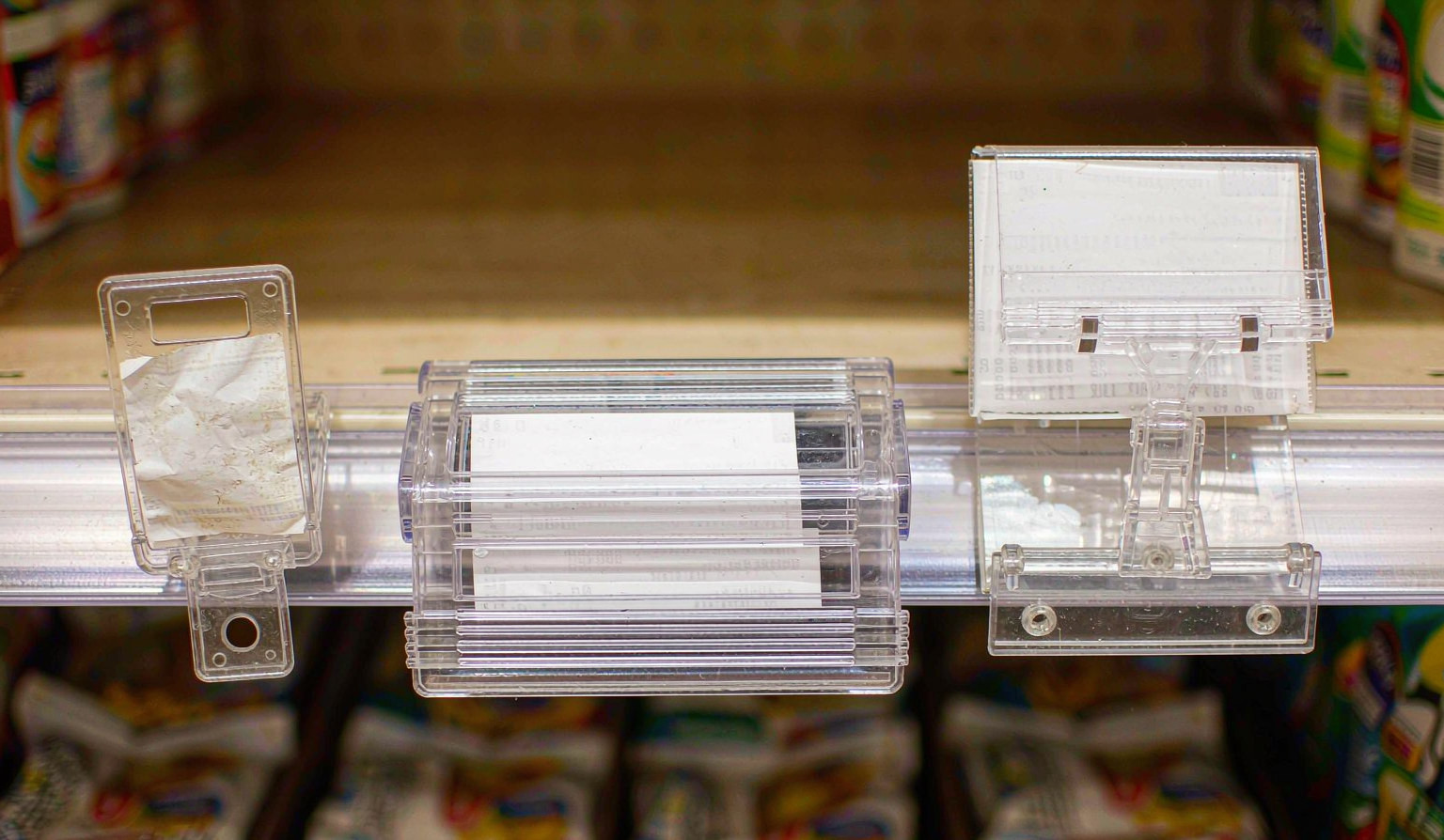 Three different clear shelf-edge price tag holders mounted in a row along one supermarket shelf lip