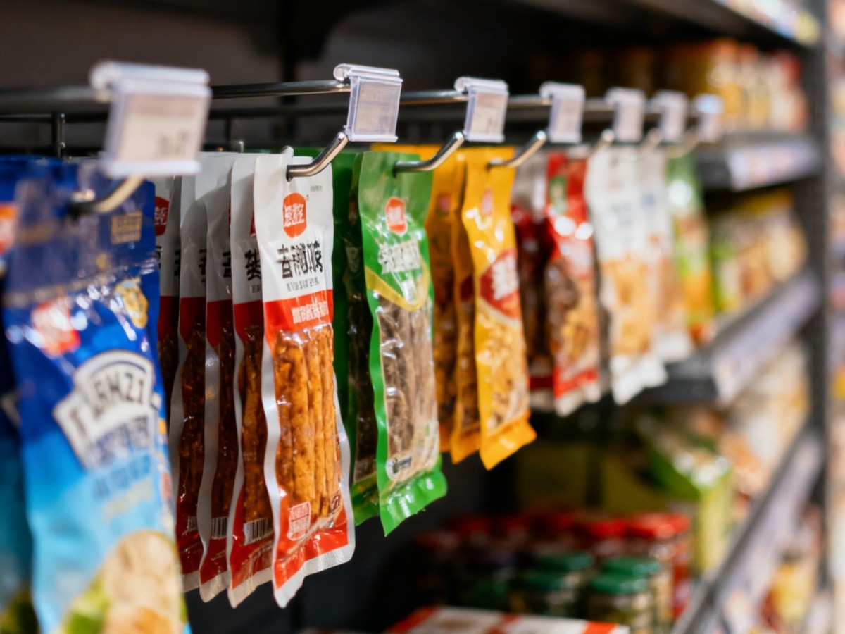 Pouched snacks hanging from single-prong display hooks with clear clip-on label holders along a supermarket shelf edge, stocked shelves behind