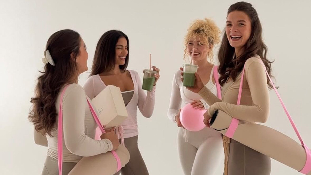 Designer and manufacturer collaborating over a detailed apparel tech pack Smiling women in neutral tone long-sleeve yoga wear and leggings holding green smoothies and rolled yoga mats.