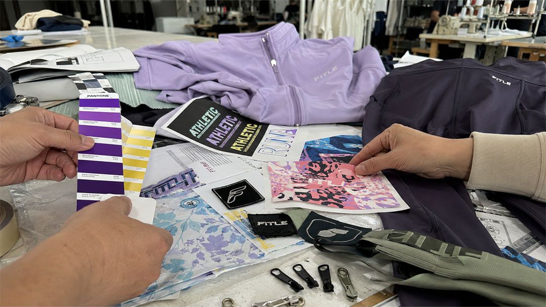 alt=A collage showing sublimation printing, embroidery, and screen printing on activewear Sportswear manufacturer reviewing custom activewear samples, Pantone color cards, printed fabric designs, logo patches, and zipper accessories on a factory worktable