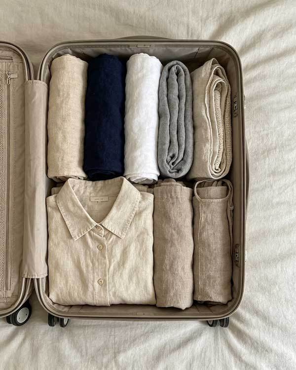 A neatly packed open suitcase showing rolled linen clothing items in neutral tones - beige, navy, white, gray - organized by color, travel packing flat lay, soft lighting, minimalist travel photography