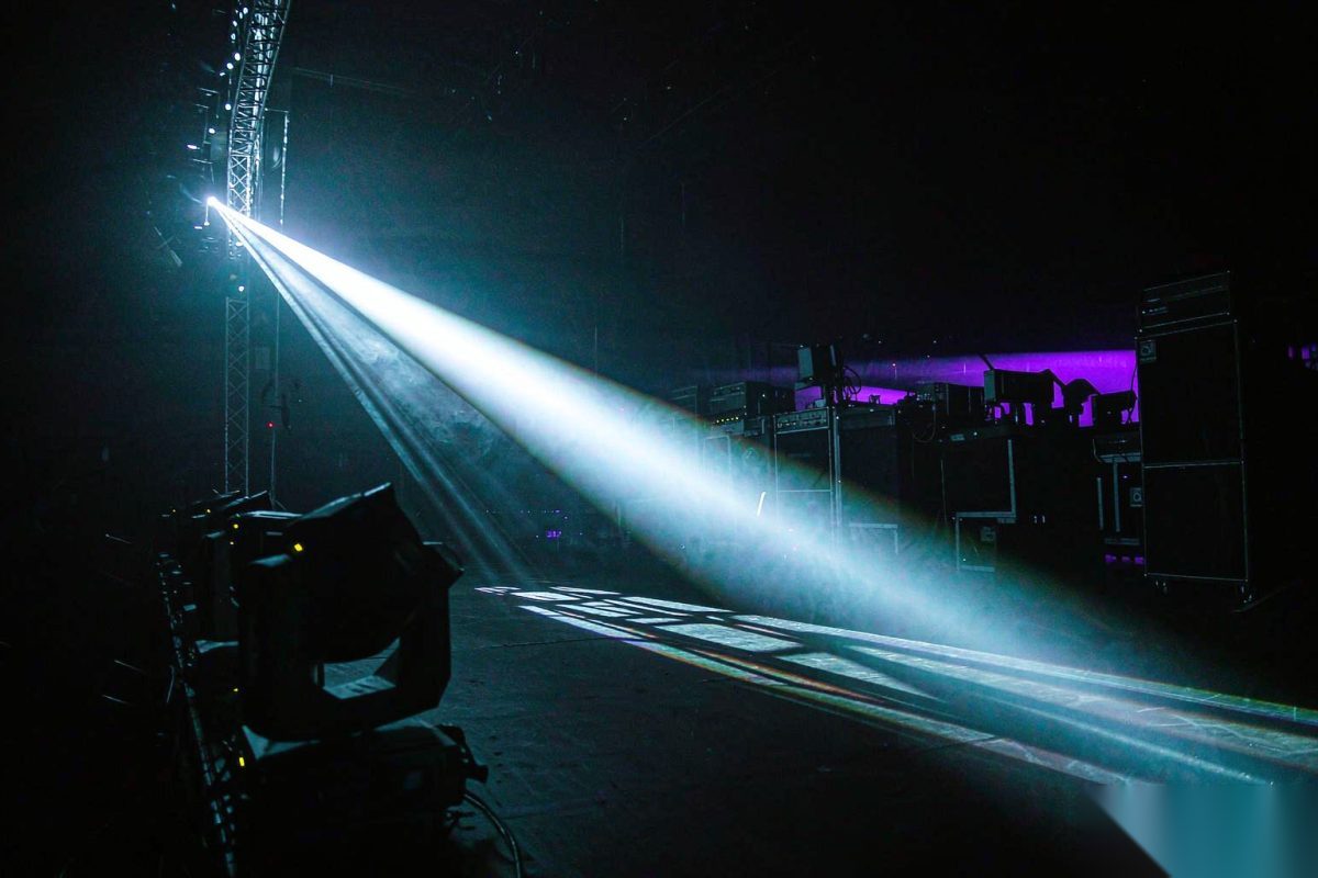 A single laser beam traveling a long throw from the rear of a dark indoor arena toward the stage, widening slightly with distance