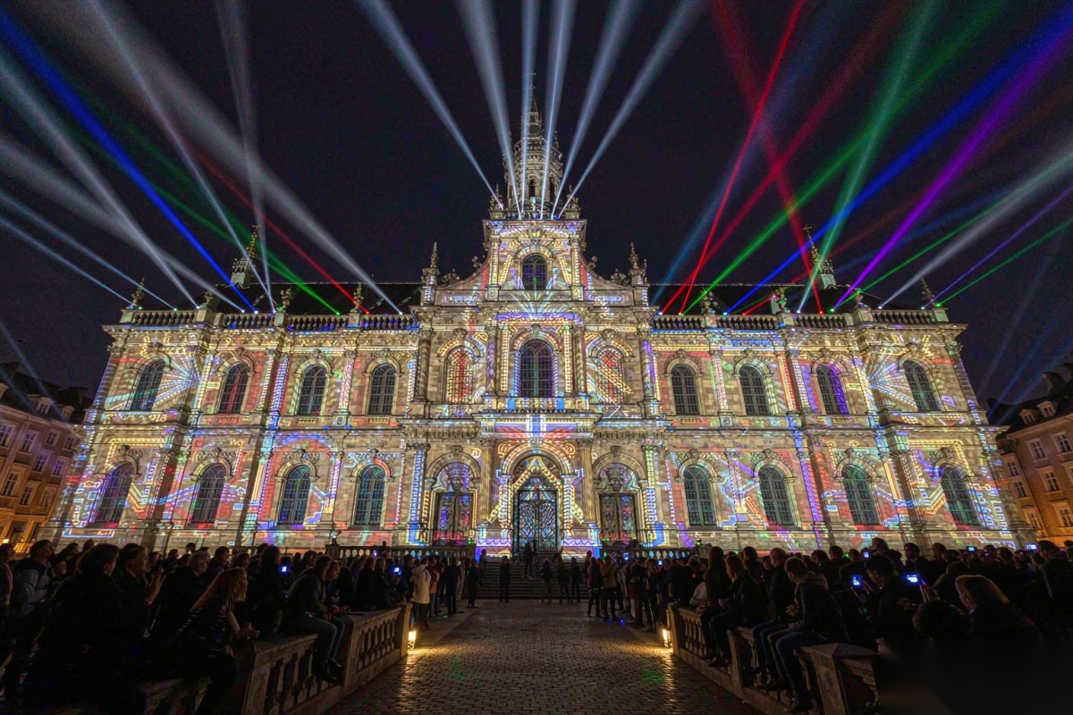 Classical building facade at a night light festival, illuminated by colorful laser projection mapping with beams fanning above the roofline