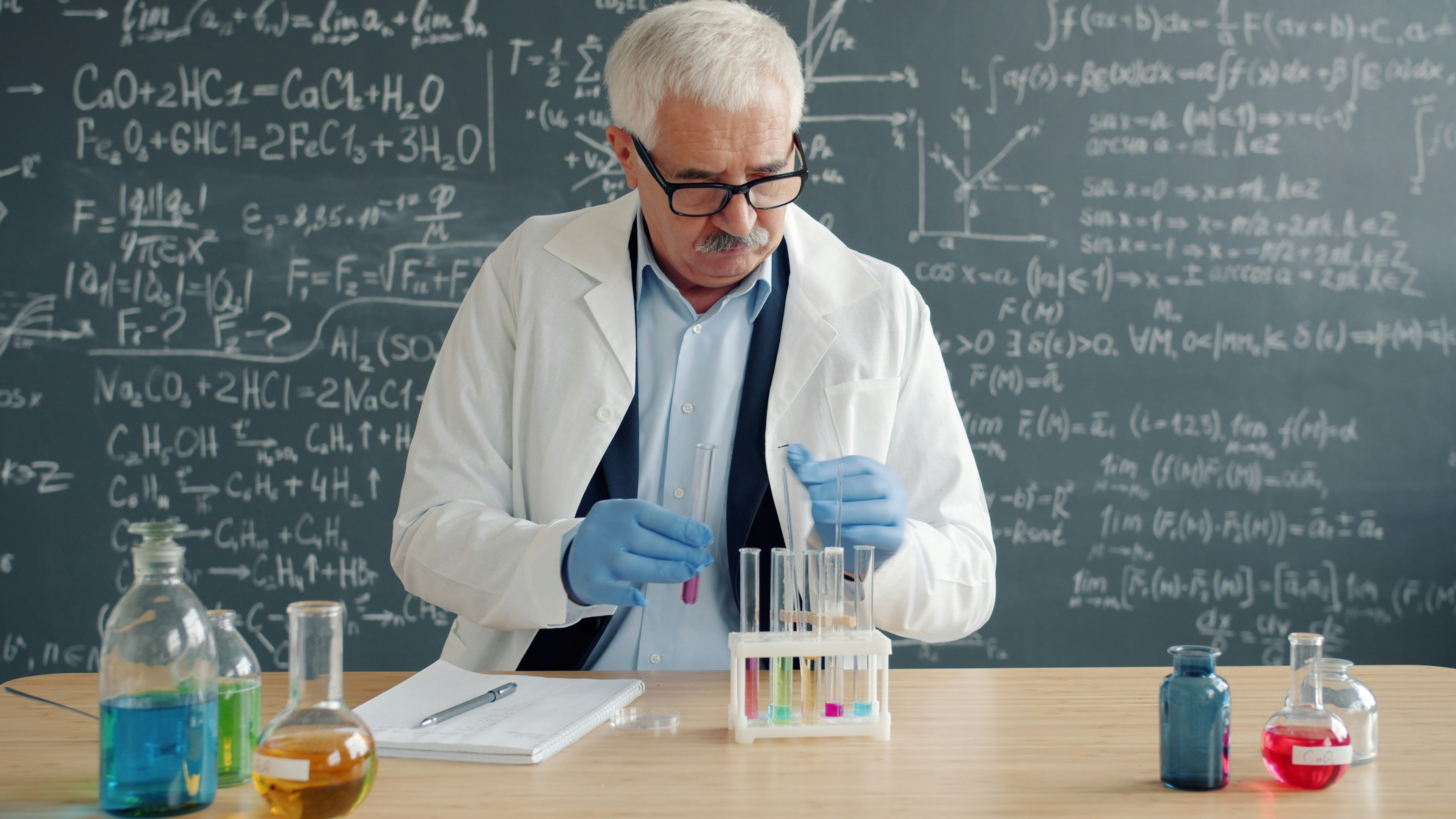 A scientist in a lab coat and safety glasses holding up a beaker for inspection in a clean, modern laboratory.
