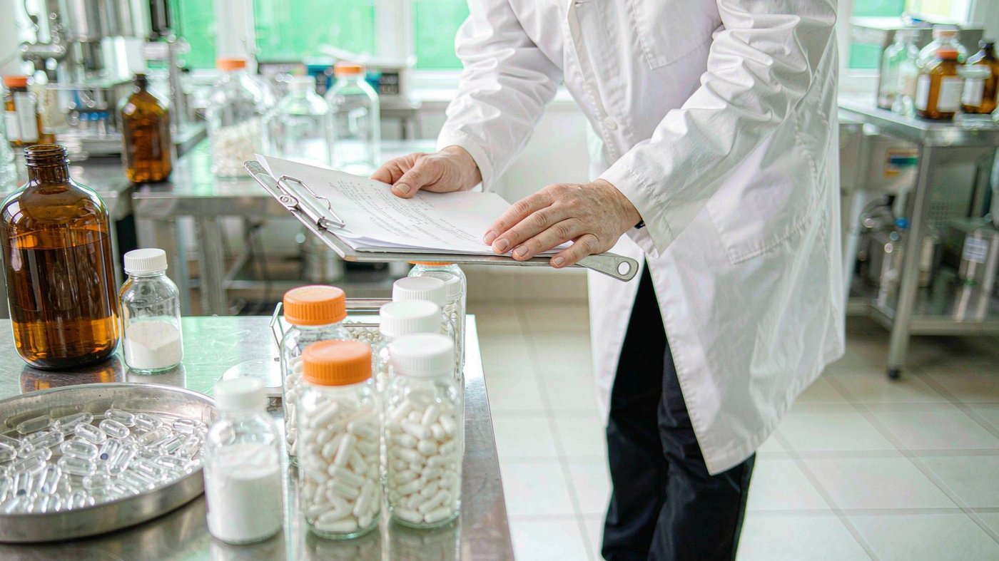 Quality assurance professional in a white lab coat reviewing a certificate of analysis beside stainless steel tables of supplement capsules and bottles