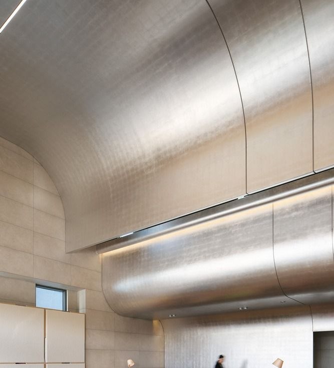 Smooth polished stainless steel ceiling plane above a reception desk, its curve catching and diffusing the ambient light