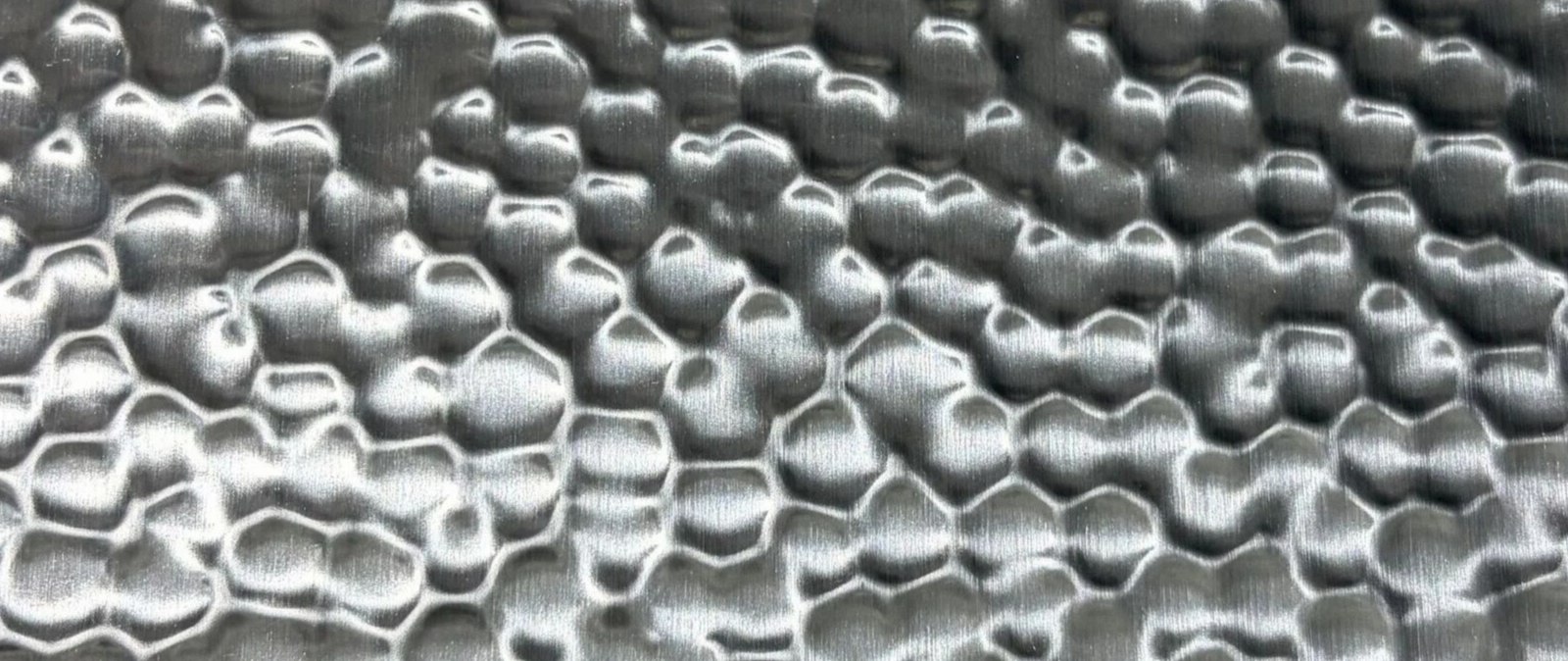 Irregular hammered relief across a natural stainless steel sheet under grazing light