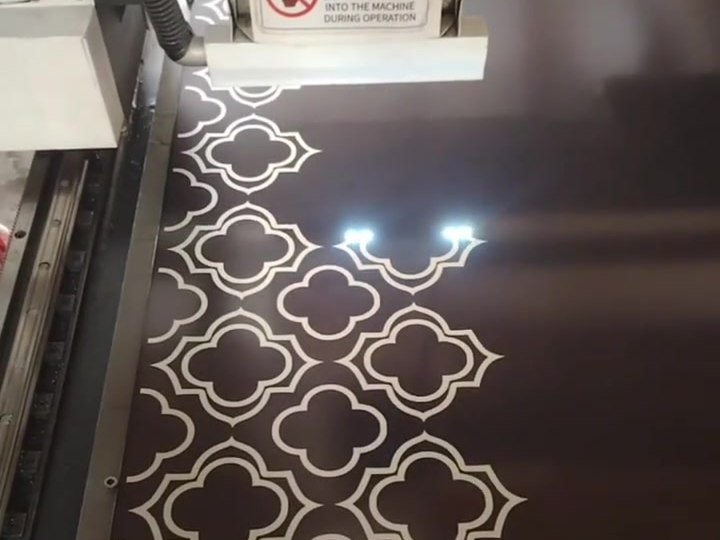 An etched quatrefoil pattern on a dark stainless steel sheet under the etching head, the etched areas reading light against the dark ground