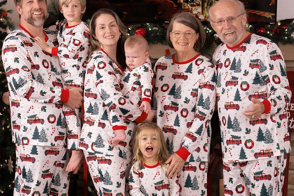 Three generations of a family wearing matching white pajamas printed with red vintage trucks, Christmas trees and candy canes, photographed beside a decorated Christmas tree