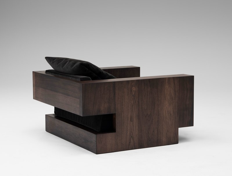 Rear three-quarter view of a sculptural walnut hotel armchair with a stepped open base and recessed velvet seat