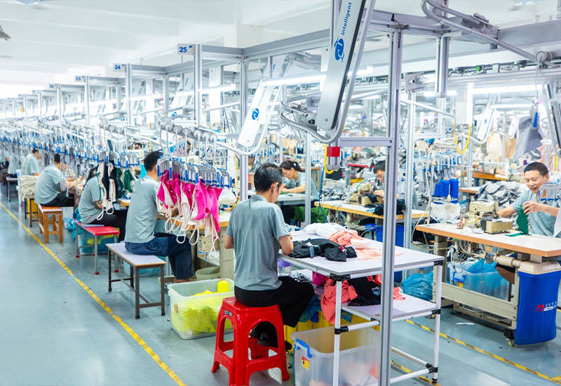 Workers producing garments on an organized apparel factory production line with sewing stations and overhead garment handling system.