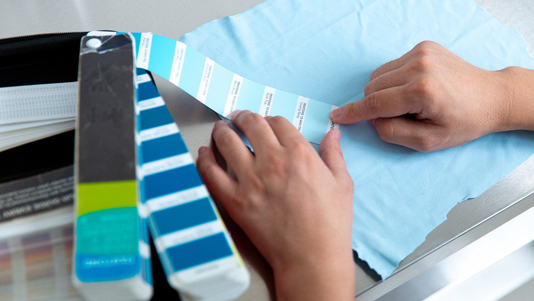Hands comparing light blue fabric with a color reference swatch book