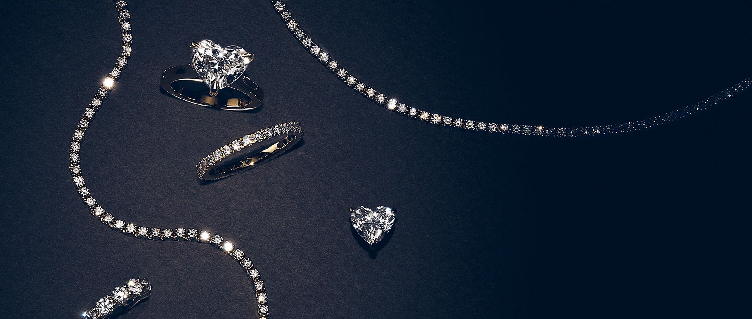 Lab-grown diamond rings and tennis chains arranged on a dark navy surface