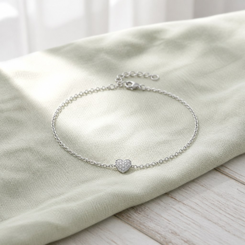 Silver heart charm bracelet laid on sage linen, fine cable chain running to the pave heart